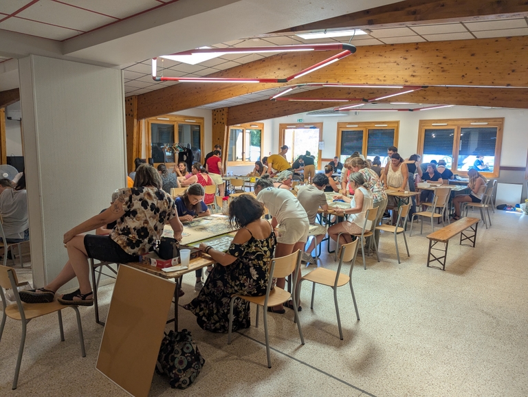 Vue de la salle des fêtes remplie de participants penchés sur plusieurs tables de puzzle